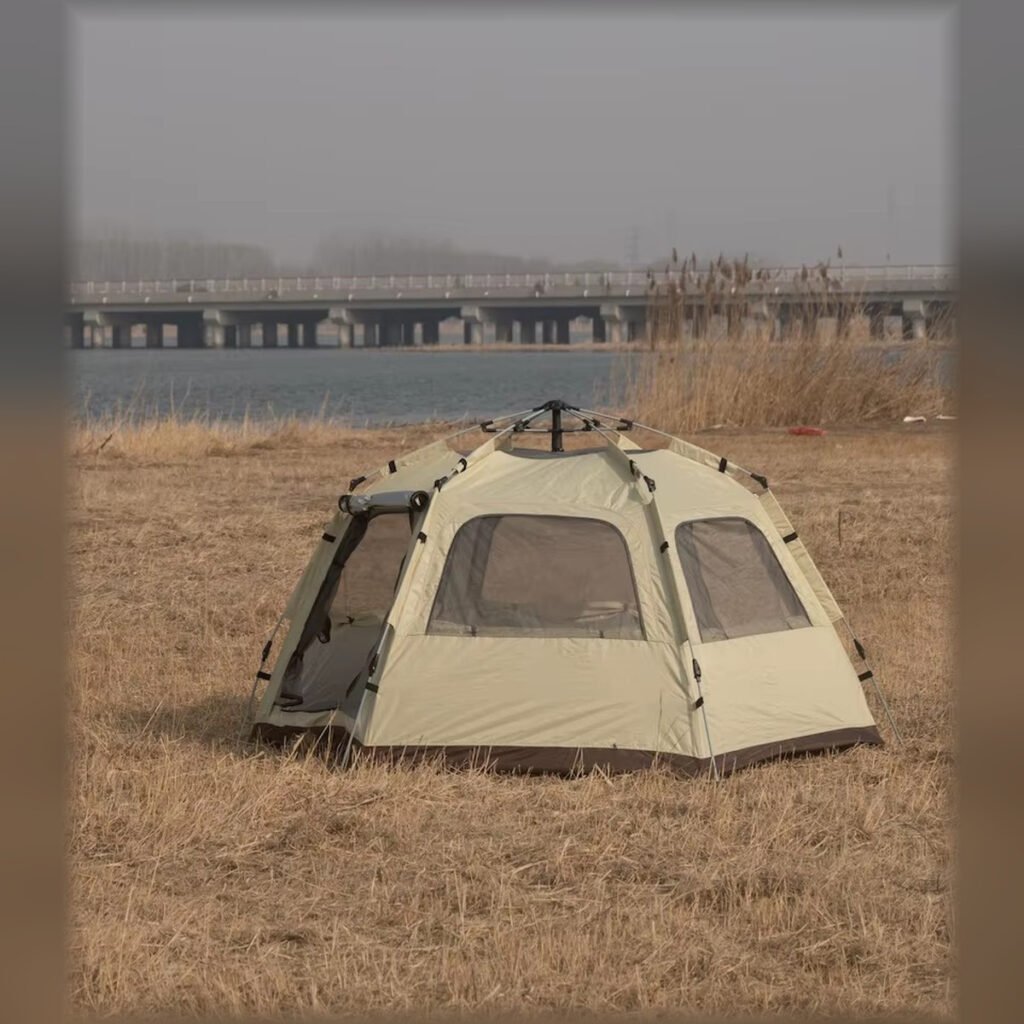 Beige hexagonal folding camping tent set up in a riverside field.