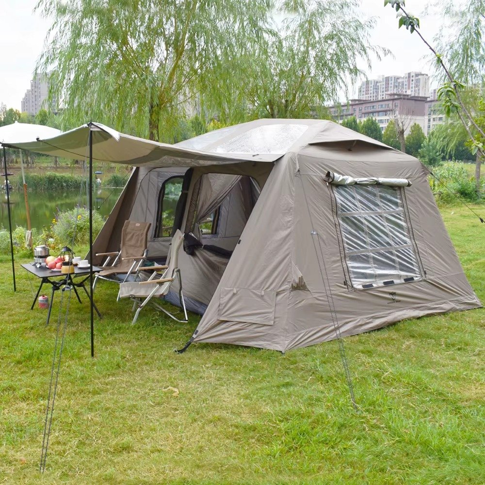 Light-colored cabin tent with an extended awning set up on grass beside a landscaped outdoor area.
